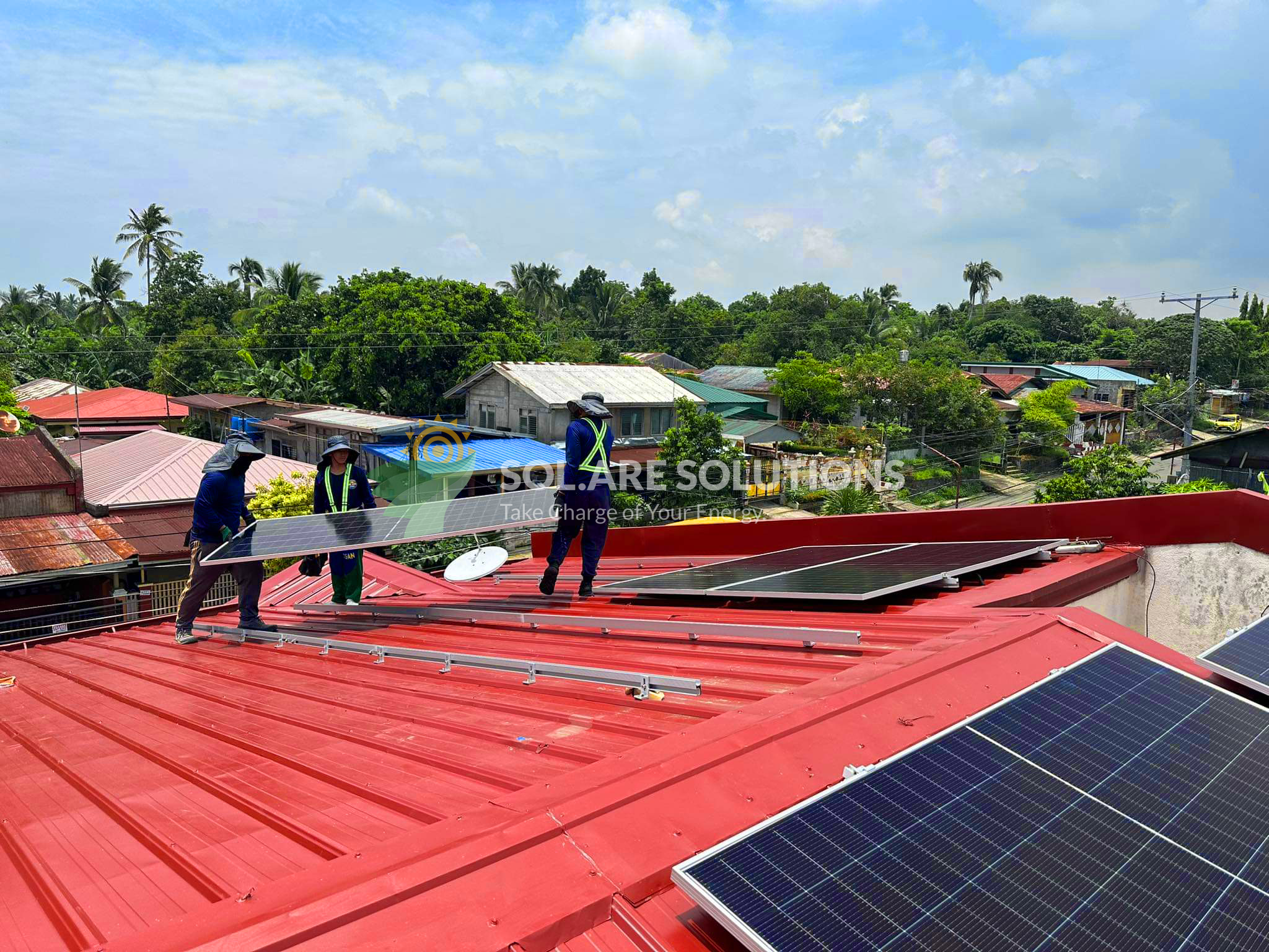 Solar installation team mounting photovoltaic panels on a red metal rooftop in a rural Philippine neighborhood, surrounded by trees and houses, under a partly cloudy sky. Branded overlay reads 'SOL.ARE SOLUTIONS – Take Charge of Your Energy
